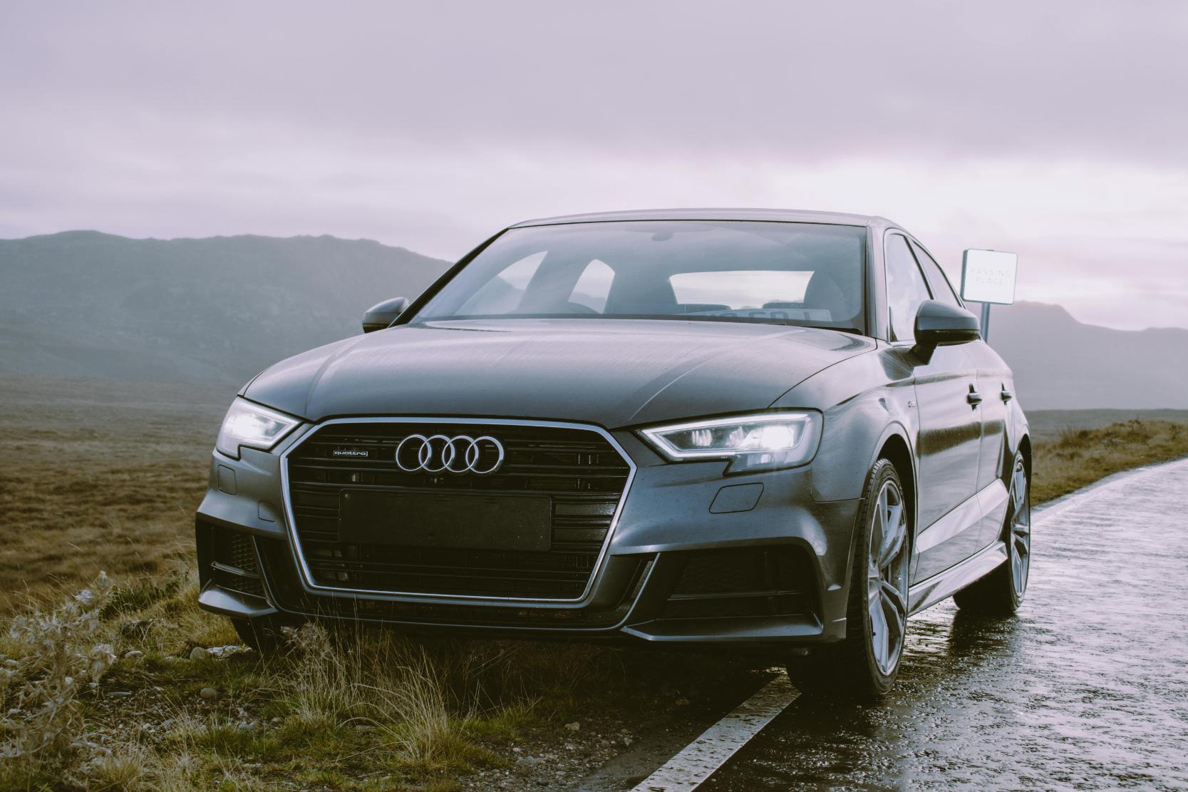 La imagen muestra un coche Audi de color gris oscuro estacionado sobre una carretera mojada en medio de un paisaje natural y montañoso.